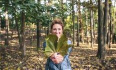 Donna sorridente che tiene foglia autunnale nella foresta — Foto stock