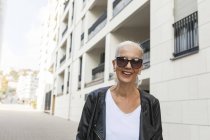 Mulher sorrindo usando óculos de sol e jaqueta de couro andando na cidade — Fotografia de Stock