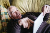 Старший чоловік, за допомогою планшетного ПК на дому в розслабленому стані, лежачи на дивані. — стокове фото
