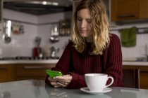 Woman sitting in the kitchen with cup of tea using cell phone — Stock Photo