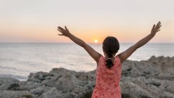 Little girl with raised arms at rocky coast at sunset — Stock Photo