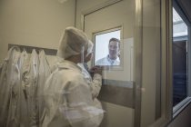 Deux techniciens de laboratoire en vêtements de protection stériles regardant un collègue derrière une vitre — Photo de stock