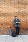 Joven guapo hipster de pie contra la pared de ladrillo con teléfono inteligente - foto de stock