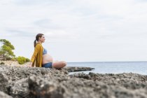 Entspannte Schwangere sitzt auf Felsen am Meer — Stockfoto
