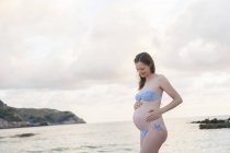 Donna incinta in piedi sulla spiaggia e la pancia toccante — Foto stock