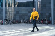 Jovem com arquivo vestindo pulôver amarelo e óculos de sol andando em frente ao prédio de escritórios — Fotografia de Stock