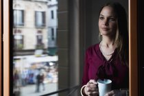 Jeune femme debout près de la fenêtre, boire du café — Photo de stock