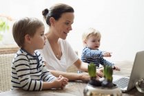 Madre usando laptop con sus hijos sentados en la mesa, vista lateral - foto de stock