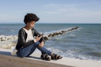 Young woman sitting by the sea, using smart phone — Stock Photo