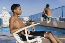 Adolescente sentado en silla piscina al aire libre mientras su freind está haciendo una inmersión de bala de cañón - foto de stock