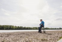 Hombre mayor sentado en el andador de ruedas a orillas del río - foto de stock