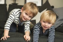 Дві маленькі хлопчики грають на дивані у себе вдома, дивлячись на камеру — стокове фото