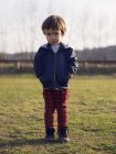 Kleiner Junge steht mit den Händen in den Taschen auf dem Gras und blickt in die Kamera — Stockfoto