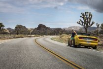 USA, California, Joshua Tree National Park, donna appoggiata alla macchina parcheggiata gialla sulla strada provinciale — Foto stock
