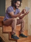 Усміхаючись молода людина, що сидить на дерев'яних сходів, дивлячись на цифровий планшетний — стокове фото