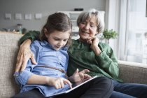 Abuela y su nieta sentadas juntas en el sofá con la tableta digital - foto de stock
