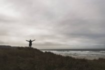 Uomo in piedi sulla costa sotto cielo nuvoloso stendendo le braccia — Foto stock
