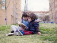 Kleines Mädchen sitzt mit ihrem Bruder auf dem Rasen und schaut sich ein Buch an — Stockfoto