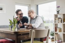 Homme âgé avec petit-fils assis à table dans le salon avec ordinateur portable et smartphone — Photo de stock