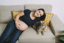 Happy pregnant woman sitting on the couch with tabby cat — Stock Photo