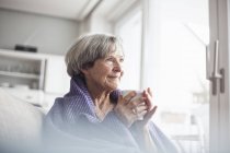 Retrato de mujer mayor feliz sentada en el sofá en casa con una taza de café - foto de stock