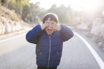 Niño gritando de pie en el carril del camino del campo vacío cubriendo los ojos con sus manos - foto de stock