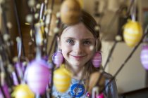 Retrato de niña sonriente detrás de ramitas de sauces de coño decoradas con huevos de Pascua - foto de stock