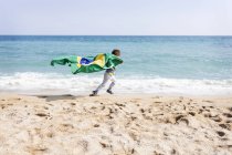 Petit garçon courant avec le drapeau brésilien sur une plage — Photo de stock
