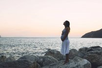 Mulher grávida em pé sobre uma rocha em frente ao mar ao pôr do sol — Fotografia de Stock