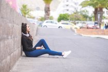 Испания, Тенерифе, улыбающаяся девочка-подросток с наушниками и смартфоном, сидящая на земле — стоковое фото