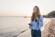 Smiling young woman lwith smartphone standing on the beach — Stock Photo