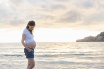 Donna incinta in piedi sulla spiaggia al tramonto — Foto stock