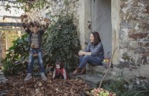 Femme assise à l'entrée de la maison de campagne et enfants jouant avec les feuilles d'automne — Photo de stock