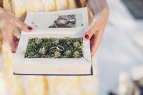 Woman's hands holding prepared book with two wedding rings lying on moss — Stock Photo