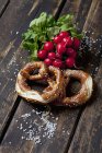 Two pretzels, red radishes and scattered salt grains on dark wood — Stock Photo