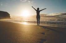 Femme debout sur la plage avec les bras levés au coucher du soleil — Photo de stock