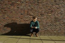Young man crouching in front of a brick wall with digital tablet — Stock Photo
