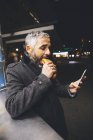 Hombre comiendo salchicha de queso Carniolan mientras mira el teléfono inteligente por la noche - foto de stock