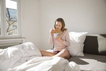 Mujer joven sentada en su cama con taza de café y croissant - foto de stock