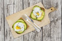 Wholemeal bread slices with sliced avocado and poached eggs on wooden board — Stock Photo