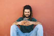 Портрет улыбающегося молодого человека с дредами и бородой, смотрящего на свой смартфон перед красноватой стеной — стоковое фото