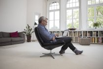 Homem maduro sentado na cadeira e usando laptop em casa — Fotografia de Stock