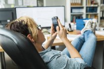 Woman reading messages on smartphone with feet on desk — Stock Photo