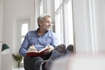 Relajado hombre maduro con libro mirando por la ventana en casa - foto de stock