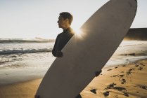 Жінка стоїть на пляжі на заході сонця і тримає дошку для серфінгу — стокове фото