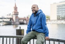 Germany, Berlin, laughing man wearing blue rainjacket sitting on railing in front of Oberbaum Bridge — Stock Photo