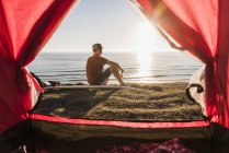 Junger Surfer campiert am Meer, sitzt am Zelt und blickt auf den Sonnenuntergang — Stockfoto