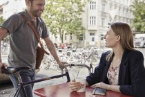 Uomo sorridente con la bicicletta che arriva in un caffè marciapiede guardando la donna — Foto stock