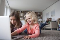 Zwei kleine Schwestern liegen mit Laptop im Wohnzimmer auf dem Boden, während ihre Eltern im Hintergrund sitzen — Stockfoto