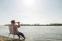 Alemania, Ludwigshafen, hombre mayor con auriculares sentado en silla plegable a orillas del río bebiendo cerveza - foto de stock